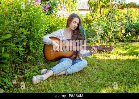 Jeune femme assise hipster dans l'herbe et jouer de la guitare sur parc ou jardin arrière-plan. Teen girl apprendre à jouer la chanson et de la musique. Passe-temps, de vie, de vous détendre, d'un instrument, l'éducation, loisirs concept Banque D'Images