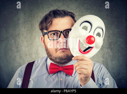 Jeune homme barbu tenant masque heureux d'être triste et sombre pendant la recherche de caméra sur fond gris Banque D'Images