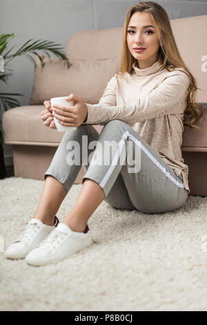 Belle jeune femme avec une tasse de boisson chaude assis sur le plancher à la maison Banque D'Images