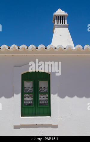 Mur Blanc d'une vieille maison avec une fenêtre avec un volet vert et un rideau. Bord du toit. Cheminée traditionnelle portugaise. Ciel bleu. Cacela Banque D'Images