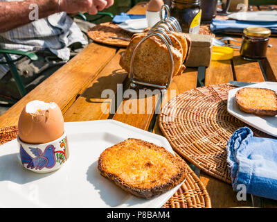 L'homme de manger le petit déjeuner en plein air en été, canicule, avec oeuf mollet et toast en rack sur Londres en bois table de jardin Banque D'Images