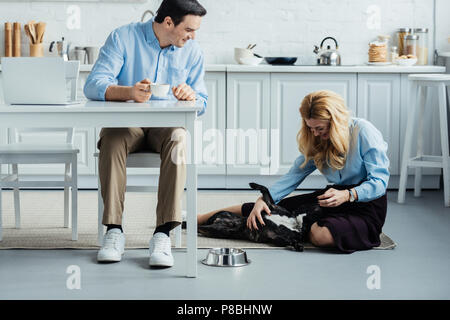 Smiling man with coffee par table avec un ordinateur portable dans la cuisine et la femme blonde caresser Frenchie dog Banque D'Images