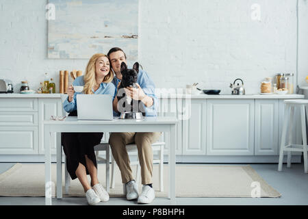 Smiling couple avec café assis par table avec un ordinateur portable dans la cuisine et de caresser Frenchie dog Banque D'Images