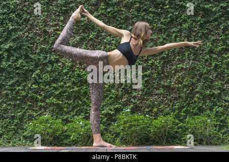 Vue de côté de belle jeune femme pratiquant le yoga en Seigneur de la danse (Natarajasana) posent devant mur recouvert avec des feuilles vertes Banque D'Images