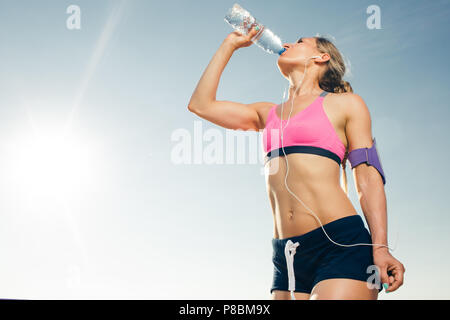 Low angle view of young de la sportive des écouteurs avec le smartphone en exécutant armband case l'eau potable de la bouteille contre le ciel bleu Banque D'Images