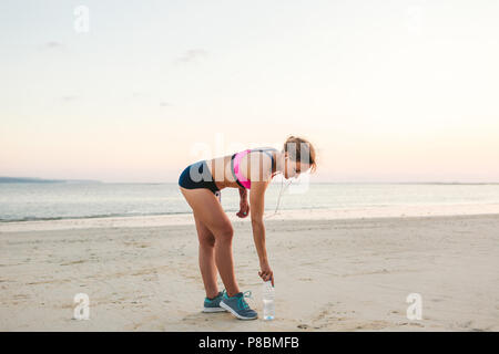 Les jeunes de la sportive des écouteurs avec brassard smartphone en cas ramasser une bouteille d'eau sur la plage avec vue sur la mer derrière Banque D'Images