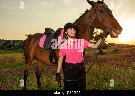 Beautiful smiling girl jockey tenir à côté de son cheval brun portant des uniformes spéciaux sur un ciel et vert sur un fond de terrain au coucher du soleil. Banque D'Images