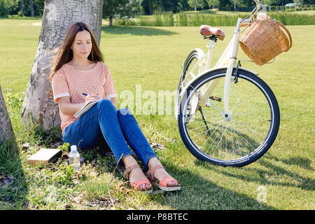 Jeune femme heureuse élève écrit dans un ordinateur portable assis sous un arbre un jour d'été Banque D'Images