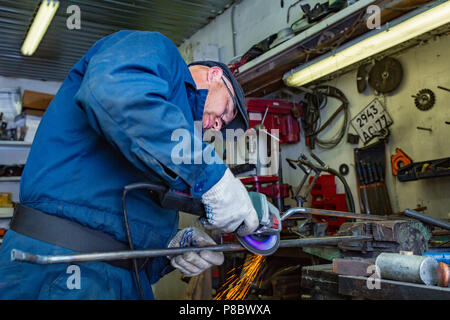 Un jeune homme au soudeur un tee-shirt bleu, lunettes et gants construction métallique processus une meuleuse d'angle dans le garage, dans l'arrière-plan beaucoup d'outils Banque D'Images