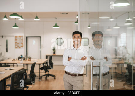 Smiling Asian businessman appuyé contre un mur de verre au travail Banque D'Images