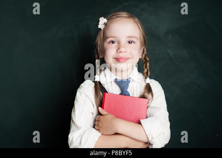 Jolie petite lycéenne enfant avec l'école livre sur fond vert tableau. Smiling in classroom élève (enfant 5-6 ans) Banque D'Images