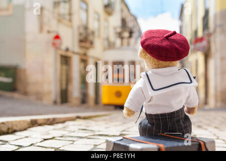 Retour de cute teddy bear wearing sailor shirt et beret hat à rue de Lisbonne (Portugal), s'asseoir sur sa valise, ancien tramway à l'arrière-plan Banque D'Images