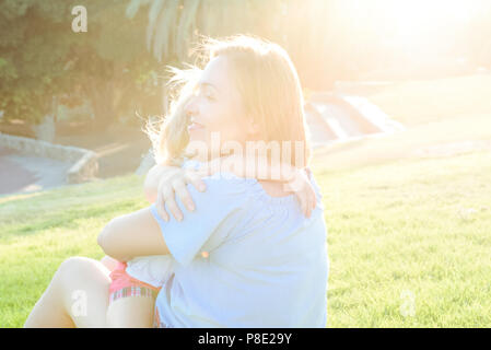 Close up mère et enfant bébé fille reposant, enlacés, rire et s'amuser sur l'herbe verte prairie au coucher du soleil. Passer du temps ensemble. Banque D'Images