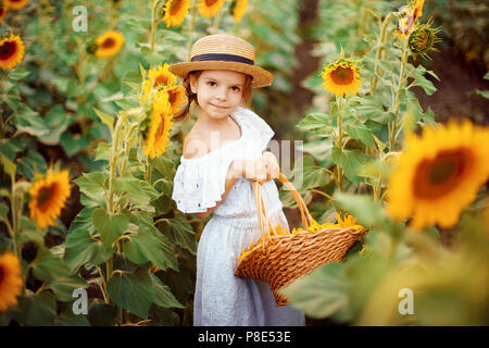 Heureux l'enfant Portrait fille en robe blanche, chapeau de paille avec un panier de tournesols en souriant et en regardant la caméra. Jeux de lumière ensoleillée onfield. Famille vie de plein air. L'humeur agréable l'été. Banque D'Images