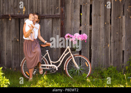 Une jeune femme brune dans une chemise blanche dans une jupe beige avec bébé sourit et s'assoit sur un vélo rétro beige avec un beau bouquet de rose p Banque D'Images