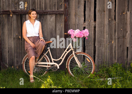 Une jeune femme brune dans une chemise blanche dans une jupe beige sourit et s'assied sur un vélo rétro beige avec un beau bouquet de pivoines rose dans un Banque D'Images