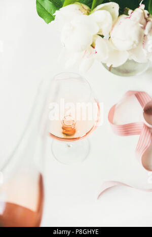 Vin rose en verre et la bouteille, rose ruban décoratif, fleurs de pivoines sur fond de table blanc, composition verticale. Célébration de l'été, mariage gr Banque D'Images