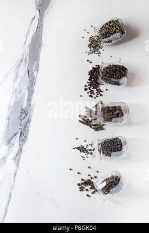 Variété de piment poivre noir, piment, poivre long, poivre des moines, le poivre et la poudre au sol dans des bocaux en verre sur la texture de marbre blanc Banque D'Images