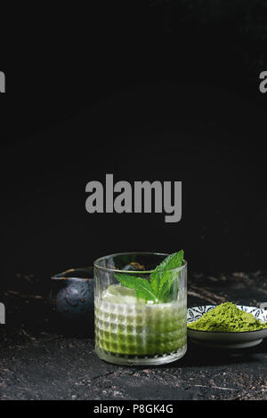 Thé vert matcha latte glacé ou un cocktail dans le verre avec des glaçons, menthe, matcha, poudre et pot de lait sur fond texture sombre Banque D'Images