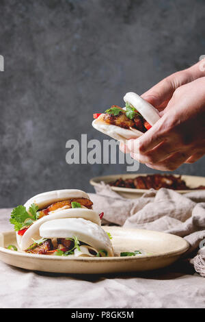 Tenir les mains de l'homme sandwich asiatique brioches bao gua à la vapeur avec du porc, du ventre et des légumes verts servi dans une plaque en céramique sur table avec nappe en lin. Comme Banque D'Images
