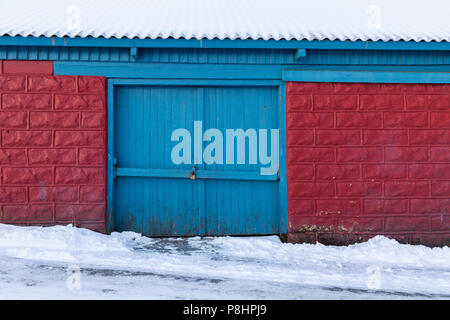Portail fermé de vieux hangar en bois, briques rouges avec porte bleue, toit et route couverte de neige en hiver Banque D'Images