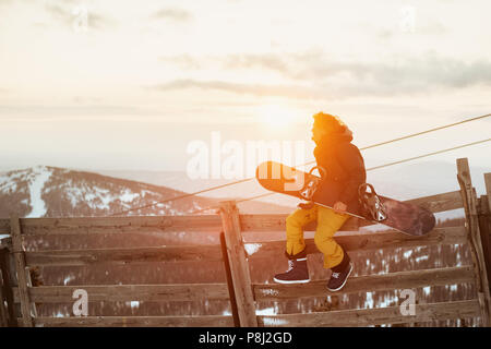 Heureux planchiste se trouve sur clôture en bois avec snowboard à mains contre sunset sky Banque D'Images