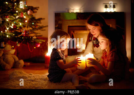 Jeune mère et ses deux petites filles assis par une cheminée tenant des bougies dans un confortable salon sombre la veille de Noël Banque D'Images