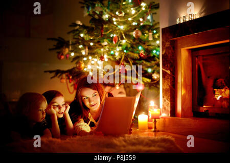 Jeune mère et ses deux petites filles de la lecture d'un livre d'un arbre de Noël dans la salle de séjour en hiver Banque D'Images