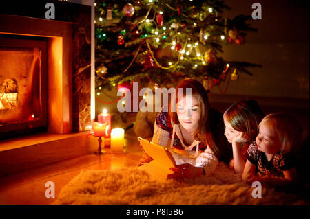 Jeune mère et ses filles à l'aide d'un tablet pc par une cheminée chaude soirée de Noël Banque D'Images