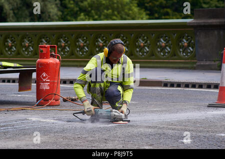 Un travailleur d'entretien des routes à l'aide d'une meuleuse d'angle power tool pour terminer la route réparations de surface. Travailleur est le port de protège-oreilles et des vis de haut. Banque D'Images