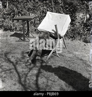 Années 1950, l'heure d'été et un homme assis à l'extérieur dans un transat a son visage et le haut du corps couvert par une grande toile carrée couvercle, vraisemblablement pour parce qu'il veut se reposer et pour protéger du soleil en personne. Il pourrait avoir joué le golf étant donné sa plus-Quatre pantalons et chaussures gofl. Banque D'Images