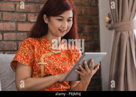 Asian woman wearing red dress sitting holding tablet sur la table dans la salle de séjour Banque D'Images