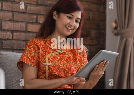 Asian woman wearing red dress sitting holding tablet sur la table dans la salle de séjour Banque D'Images