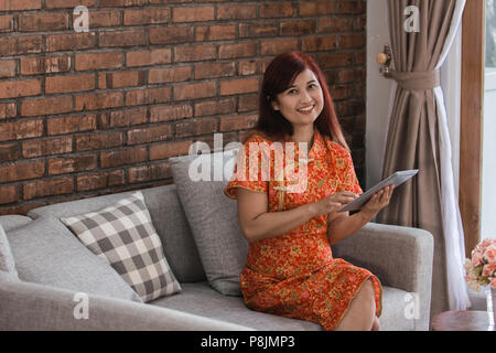 Attractive Woman wearing red dress sitting holding tablet sur la table dans la salle de séjour Banque D'Images