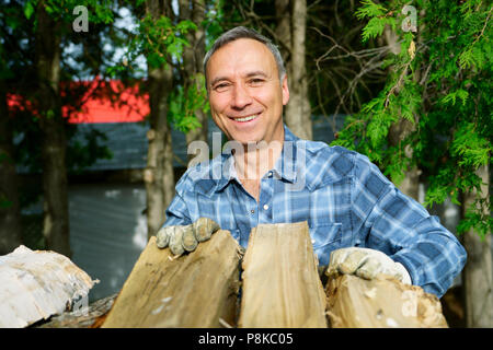 Un 50 ans caucasien homme portant une chemise bleu sourit à nous pendant que ses pieux de bois pour la saison d'hiver, pour chauffer la maison. Banque D'Images