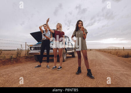 Trois jeunes femme bloqués en plein milieu d'une route de campagne avec ventilées voiture en utilisant leur téléphone pour appeler l'aide d'urgence. Banque D'Images