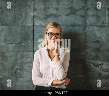 Portrait de la belle jeune femme en tenue de cérémonie se tenant contre le mur gris et souriant. Woman with glasses looking at camera with hand on chi Banque D'Images