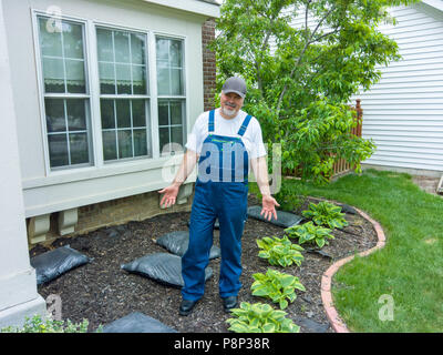 Homme debout, dans un parterre de fleurs avec des sacs de paillis faisant des gestes avec ses mains pour montrer qu'il est temps de faire le printemps le paillage autour de ses plantes Banque D'Images