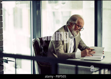 Close up side view photo de grisonnant mature businessman travaillant dans le bureau moderne avec fenêtre panoramique. Lisez le livre avec stylo. souligner interestin Banque D'Images