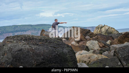 Homme était debout sur des rochers sur la plage de Ravenscar pointant vers la mer et de joints qui sont hors de l'appareil photo. Banque D'Images