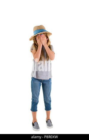 Petite fille en larmes dans les jeans et un chapeau de paille couvrant ses yeux avec ses mains comme elle se tient en pleurant isolated on white Banque D'Images