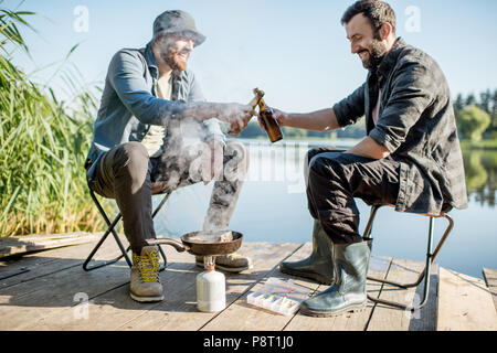 Deux pêcheurs de poissons friture assis avec de la bière pendant le pique-nique sur la jetée en bois près du lac, dans le matin Banque D'Images