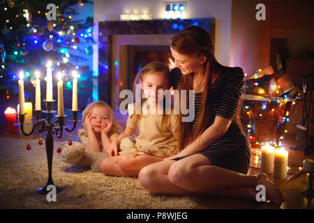 Jeune mère et ses deux petites filles assis par une cheminée dans un confortable salon sombre la veille de Noël Banque D'Images