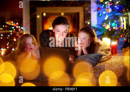 Jeune mère et ses filles à l'aide d'un tablet pc par une cheminée chaude soirée de Noël Banque D'Images