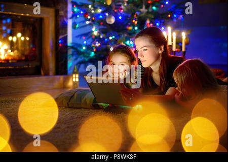 Jeune mère et ses filles à l'aide d'un tablet pc par une cheminée chaude soirée de Noël Banque D'Images