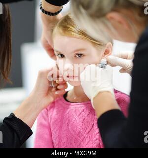 Adorable petite fille ayant le perçage des oreilles processus avec équipement spécial dans le centre de beauté par un professionnel de la santé Banque D'Images