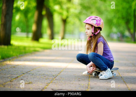 Cute little preteen girl wearing helmet assis sur une planche à roulettes dans le magnifique parc d'été Banque D'Images