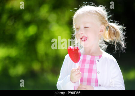 Cute little girl eating énorme sucette en forme de cœur à l'extérieur sur beau jour d'été Banque D'Images