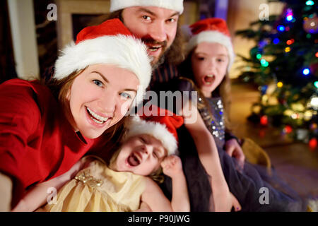 Famille de quatre jeunes heureux de prendre une photo d'eux-mêmes par une cheminée dans un confortable salon sombre la veille de Noël Banque D'Images