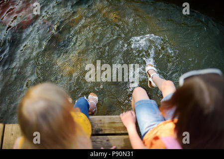 Deux mignonnes petites filles assis sur une plate-forme de bois par la rivière ou lac trempant leurs pieds dans l'eau chaude journée d'été sur Banque D'Images